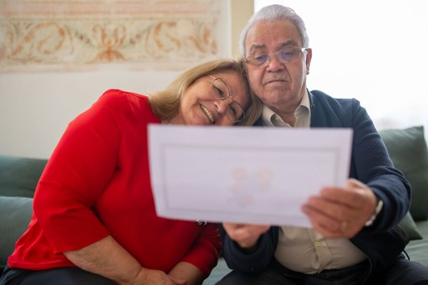 Thoughtful senior couple sits at a bright kitchen table, reviewing financial papers, representing the stress of managing h...