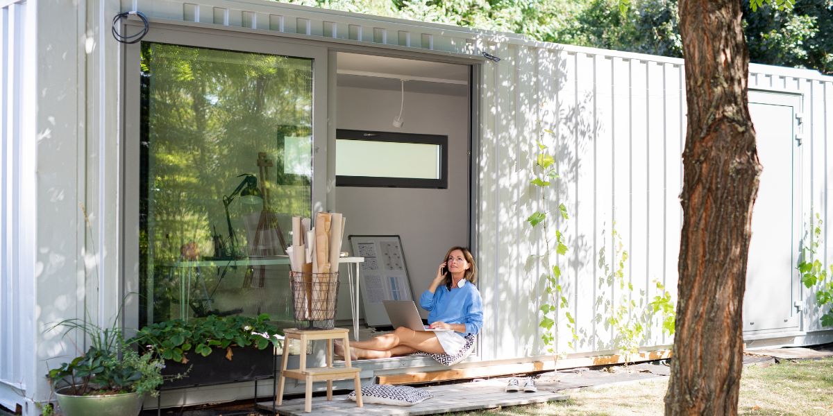 Woman sitting outside a modern container home, working on a laptop while talking on the phone.