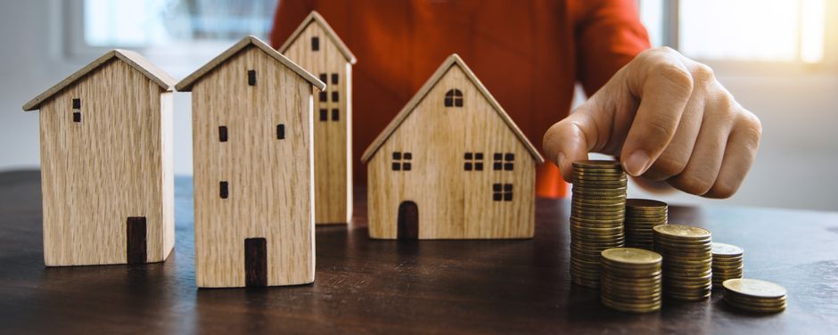 Clean, minimalist photo of a small white model house resting on a stack of coins against a bright, neutral background, sym...