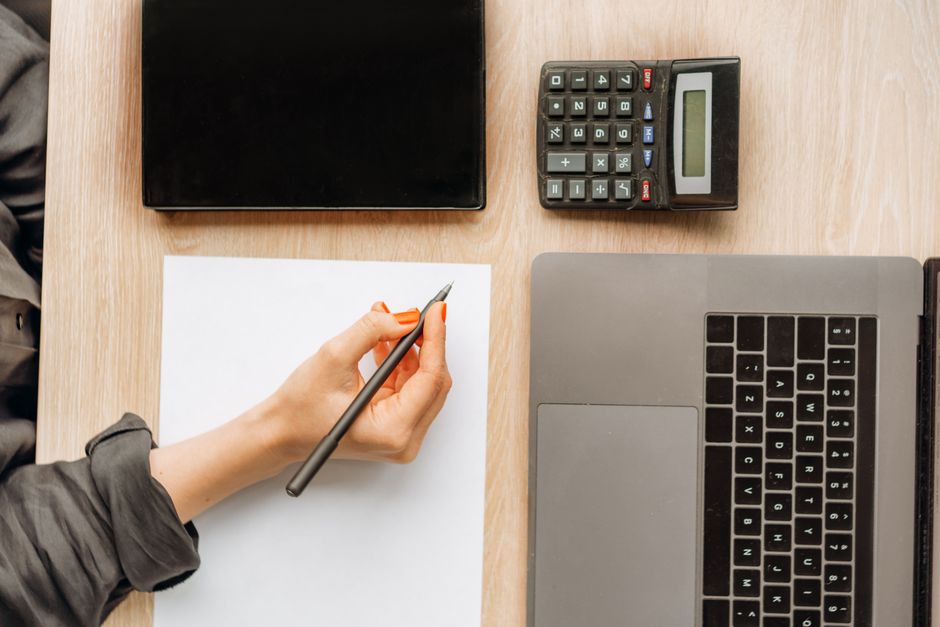 An overhead view of a hand with a calculator hovering over architectural blueprints, symbolizing the calculation of excessive real estate commissions.