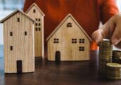 Clean, minimalist photo of a small white model house resting on a stack of coins against a bright, neutral background, sym...